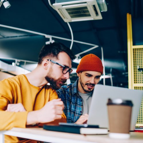 Skilled male it developers concentrated on programming cooperating while updating software on laptop computer, hipster guys studying together watching webinar on educational website of college
